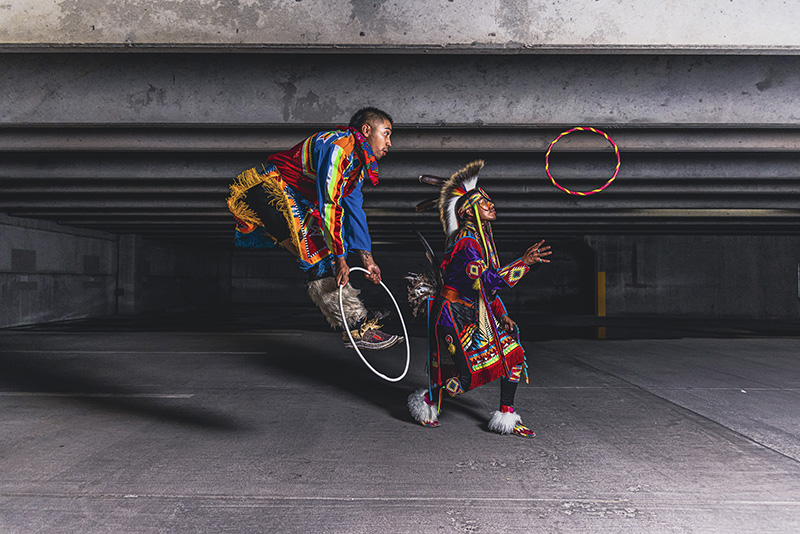 Two members of dance troupe Indigenous Enterprise in colorful regalia against a concrete backdrop, one jumping through a hoop. 