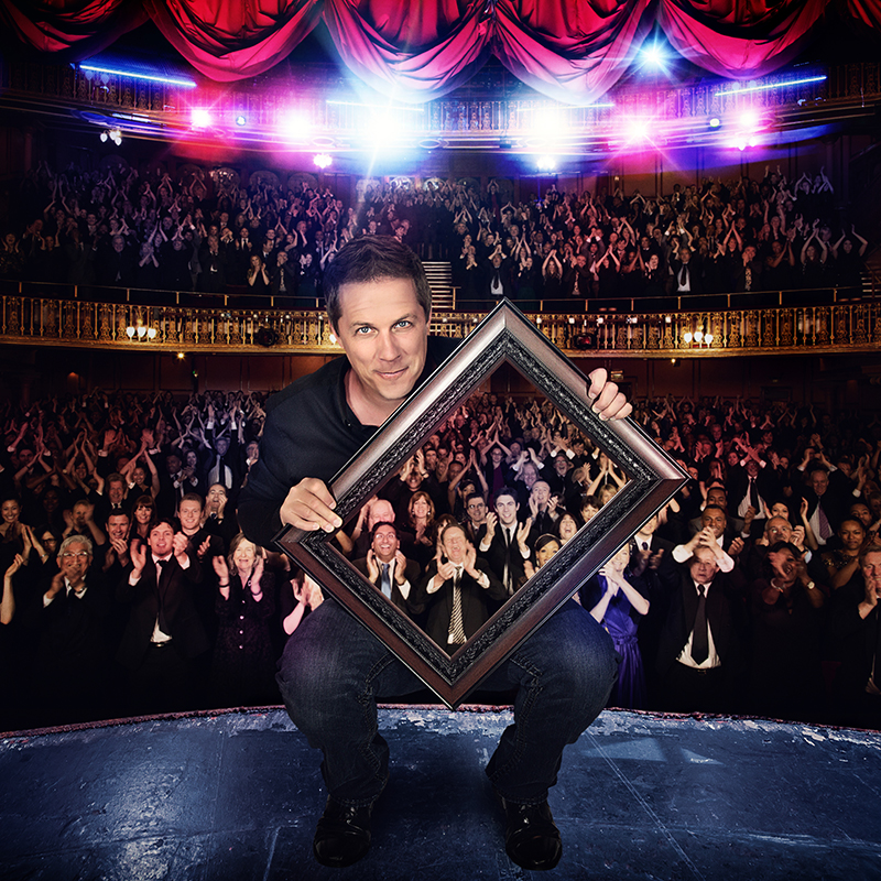 Illusionist Mike Super hold a large picture frame in front of his body, revealing hundreds of audience members sitting behind him.