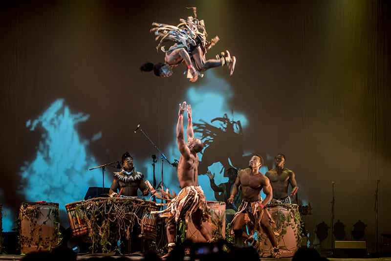 Shirtless male performers in African dress look on as an acrobat prepares to catch another acrobat somersaulting through the air.