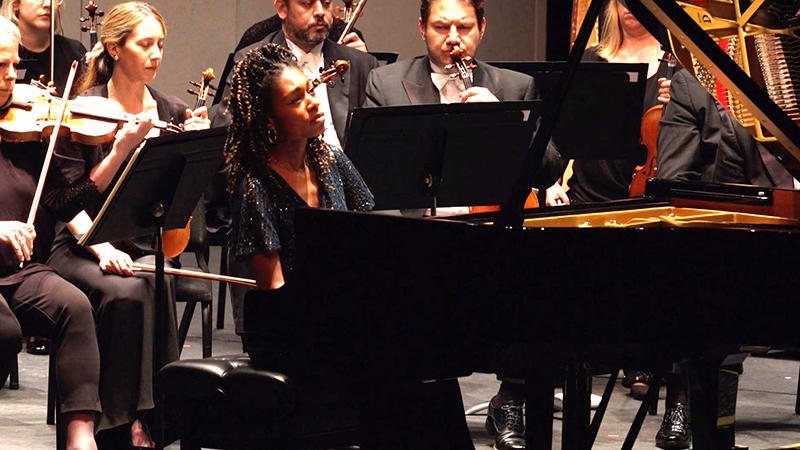 Pianist Isata Kanneh-Mason sits at a grand piano onstage, backed by the Royal Philharmonic Orchestra.