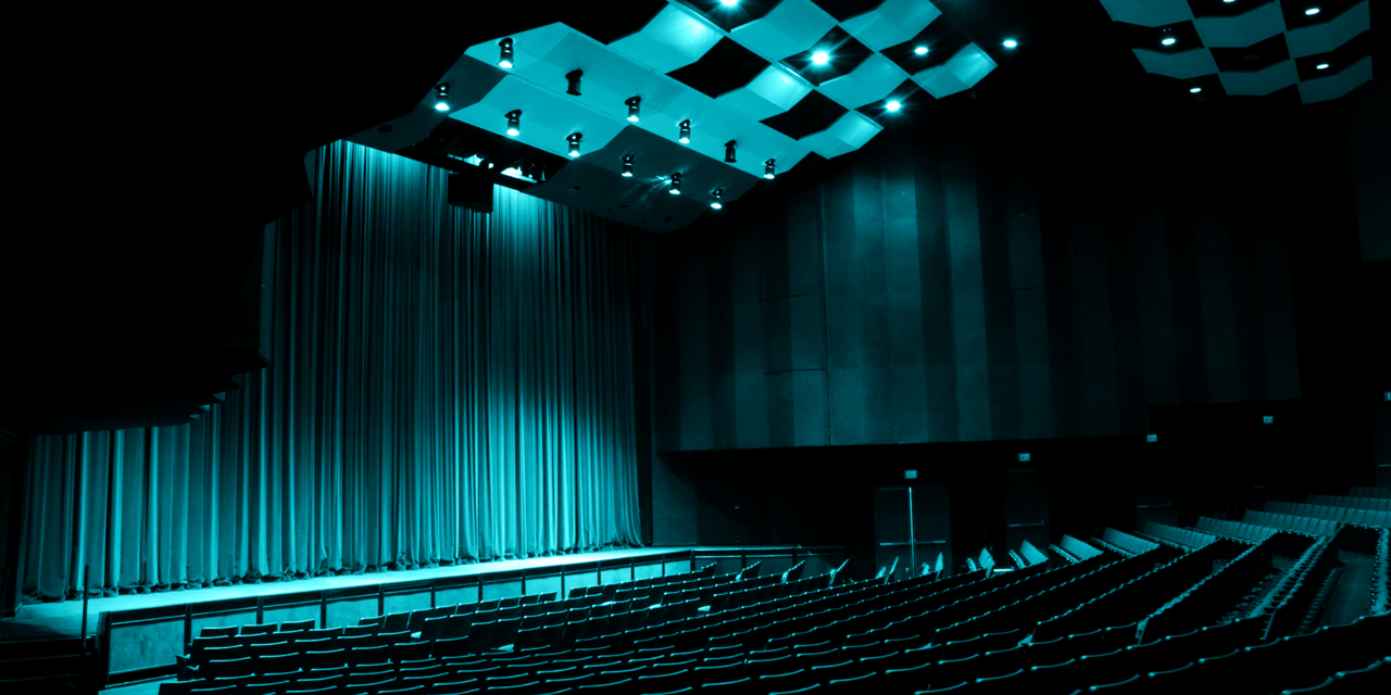 An empty stage in a performing arts space, with theatrical seating under blue lighting.
