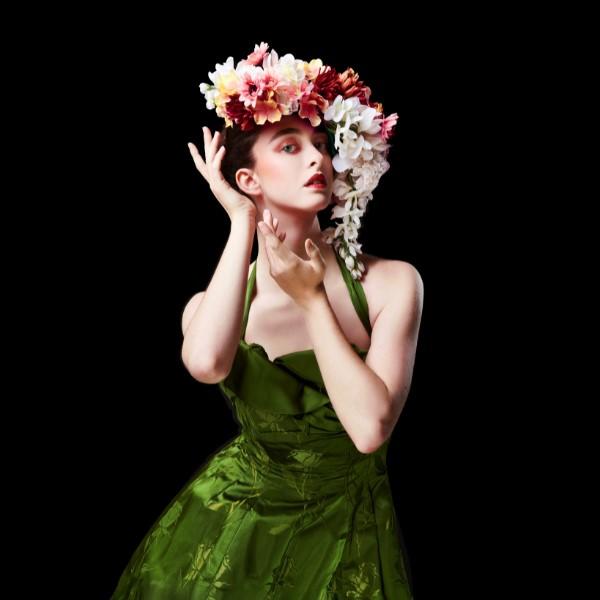 White woman with brown hair wearing a green silk off shoulder dress with big bouquet of flowers in hair against a black background