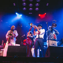 Members of the Preservation Hall Jazz Band from New Orleans perform on saxophone, trumpet, trombone, and upright bass onstage behind large speakers.