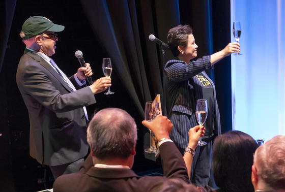 Man wearing baseball cap raises glass with woman with dark short hair towards a crowd.