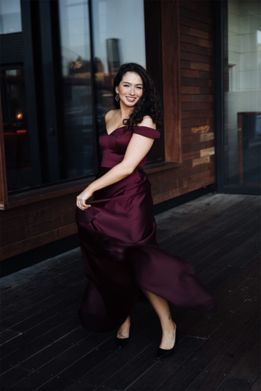 Mezzo-soprano Lisa Marie Rogali, a woman with long curly dark brown hair, wears a long burgundy, off-the-shoulder dress, standing in front of a glass building's exterior.