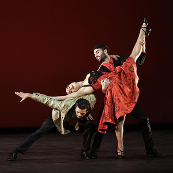 A female dancer leans back, right arm fully extended and kicking left leg in the air as a male dancer on her right, bent at the waist supports her torso and extended arm and a male dancer on her left supports the extended leg.