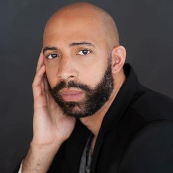 Composer Damien Geter, a bearded black man in a black shirt, rests his face in his right hand and looks at the camera.