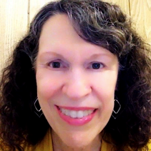 The photo shows a woman having fair skin with rosy undertones, a long oval facial shape, hazel eyes, and thick wavy shoulder-length dark brown hair lightly streaked with gray. It is parted on the left with long diagonally curving side bangs at right. A metallic drop earring of concentric black and gold diamond outline shapes dangles by and below each cheek as she smiles. Her golden yellow cardigan top contrasts with her dark hair, also in high relief against a tan wood-paneled back wall.