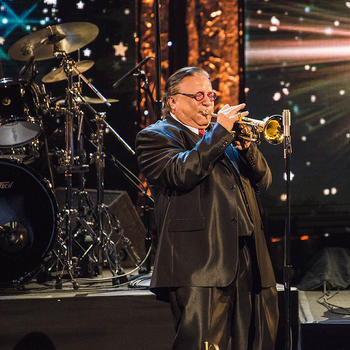 Arturo Sandoval, wearing red glasses and a blue suit, plays the trumpet onstage in front of a drumset.