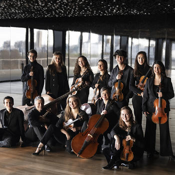 Members of the French Baroque ensemble Les Arts Florissants pose in front of a window showing a city skyline—in two rows holding period instruments, one row seated, one standing.