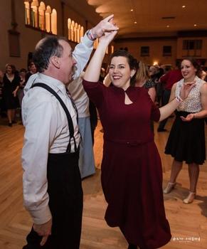 A man at left wearing a white shirt, black pants, and black suspenders, twirls his smiling female dance partner, wearing a burgundy dress.